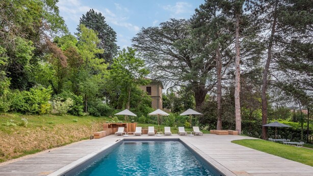 A rectangular swimming pool with a wooden deck, surrounded by lounge chairs and umbrellas, set in a lush garden with tall trees and a building in the background. - La clarté - wonderful refurbish home with garden and pool (Champagne-au-Mont-d'Or)