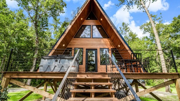 Eine Holzhütte mit einem Giebeldach, großen Fenstern und einer Holzterrasse mit Treppen, die zu einem Balkon führen.