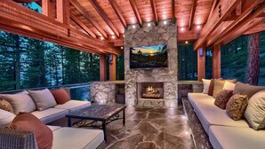 A covered outdoor seating area with stone flooring, a stone fireplace, and a TV mounted on the wall. - 450 Jill Court | 5BR/5.5BA Incline Village, Lake Views, Hot Tub (Incline Village)