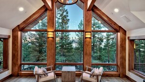 A spacious room with large windows offering a view of a forest. There are two chairs and a small table in the center. - 450 Jill Court | 5BR/5.5BA Incline Village, Lake Views, Hot Tub (Incline Village)