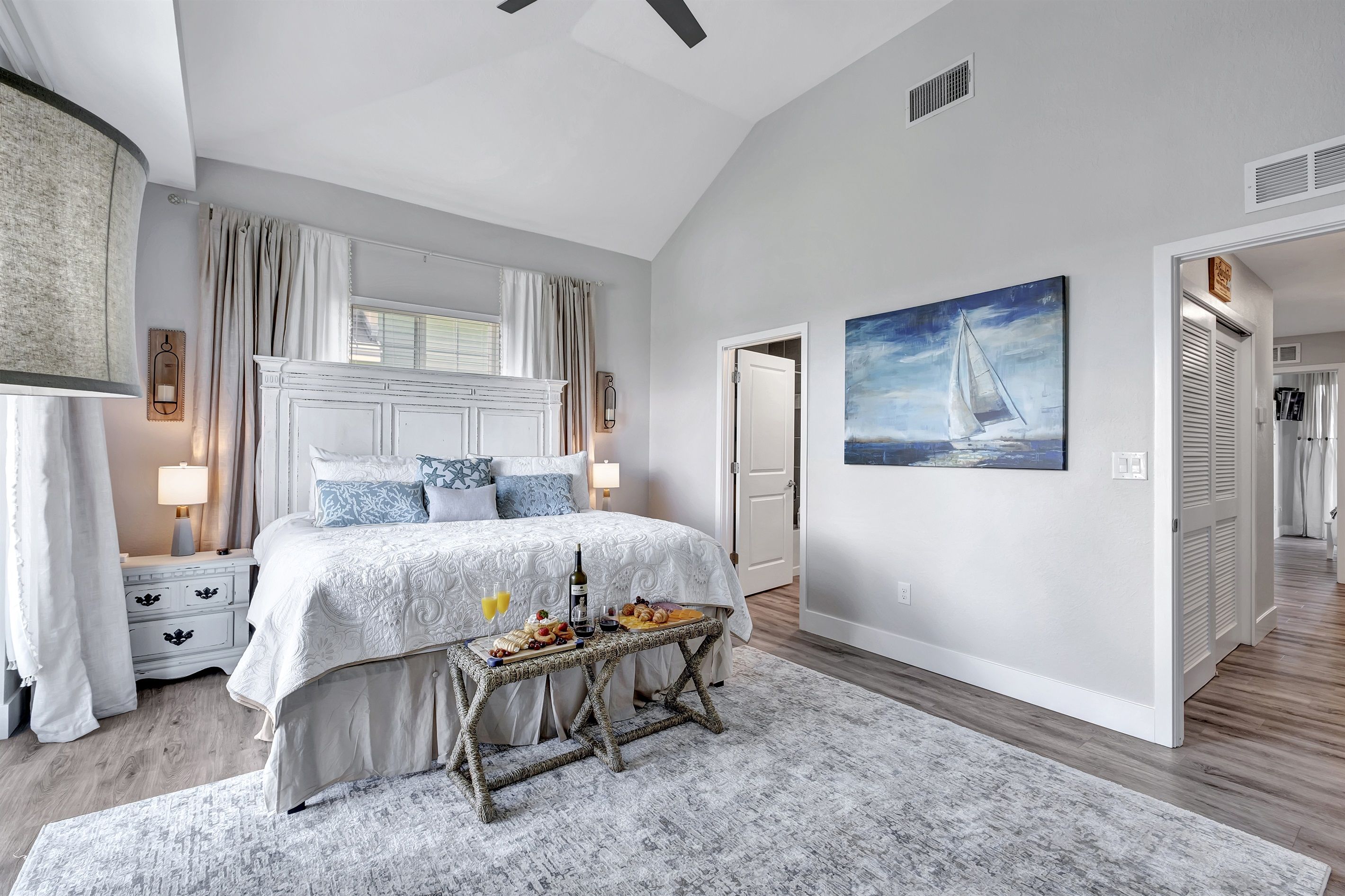 Un dormitorio con una cama grande, mesitas de noche, una lámpara de noche, una ventana con cortinas, un cuadro y una puerta que da a otra habitación.