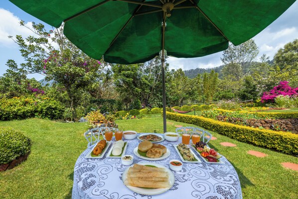 Un espacio de comedor al aire libre con una mesa cubierta por un paraguas verde, rodeado de abundante follaje y un jardín cuidado.