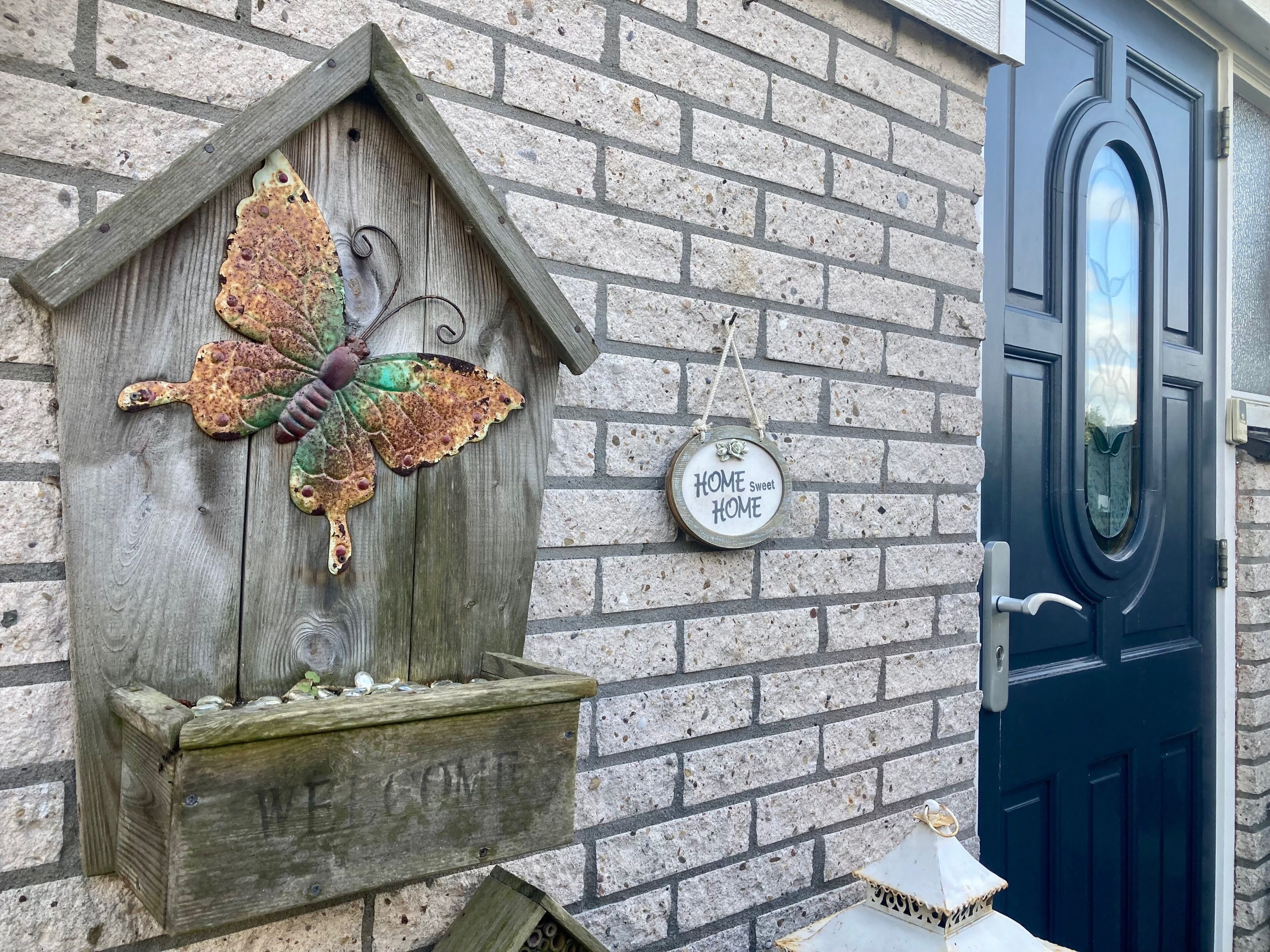Une nichée à oiseaux en bois, décorée d’un motif de papillon, avec une pancarte indiquant « Chez soi, c’est le plus beau » et une lanterne fixée à un mur de briques.