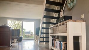 A spacious room with a wooden floor, a staircase, a bookshelf, and a window with a view of the outside.