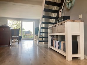 A spacious room with a wooden floor, a staircase, a bookshelf, and a window with a view of the outside.
