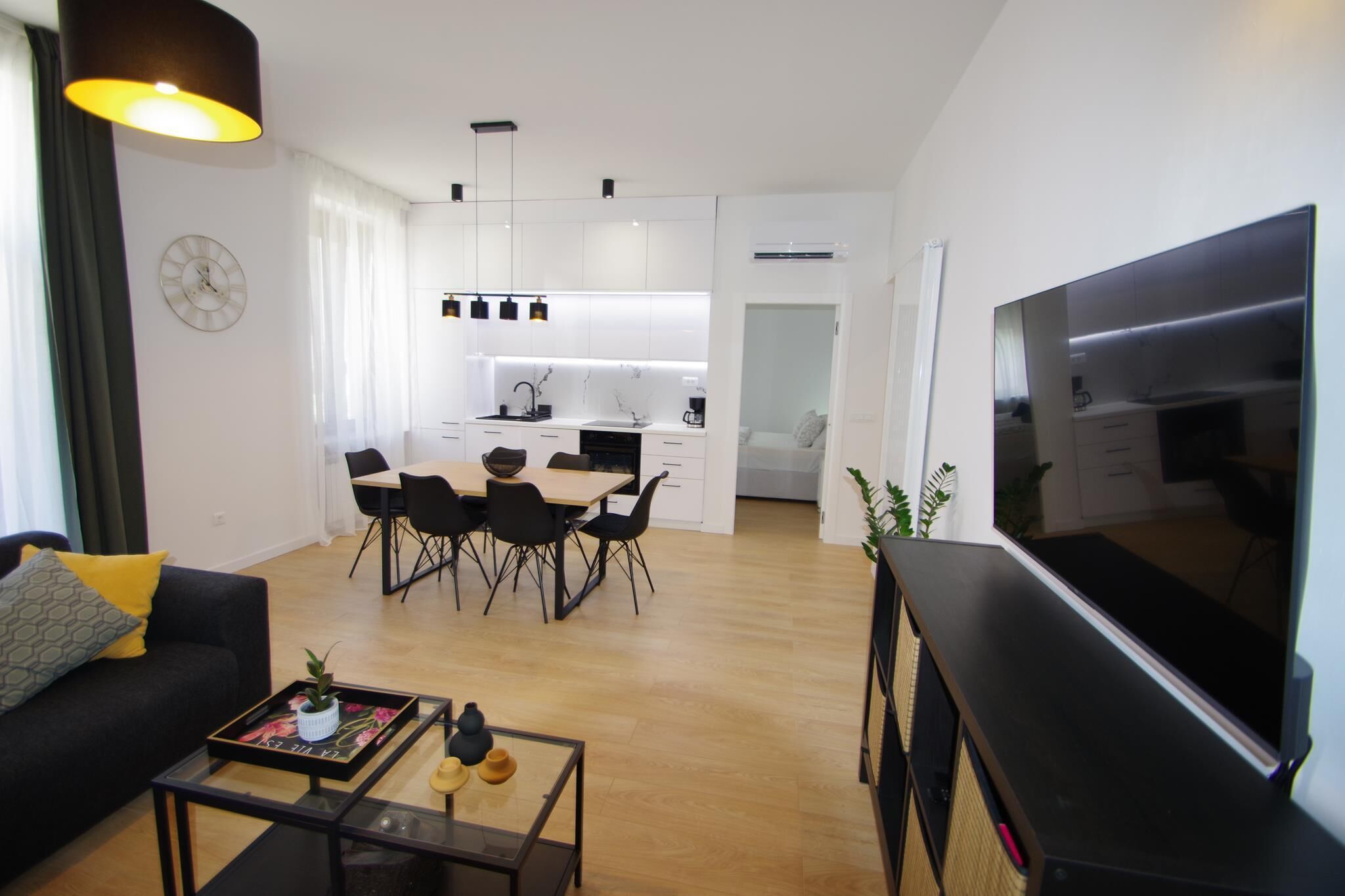 Una sala de estar moderna con un sofá, una mesa de centro y un televisor. Se ve una cocina con armarios blancos y un comedor al fondo.