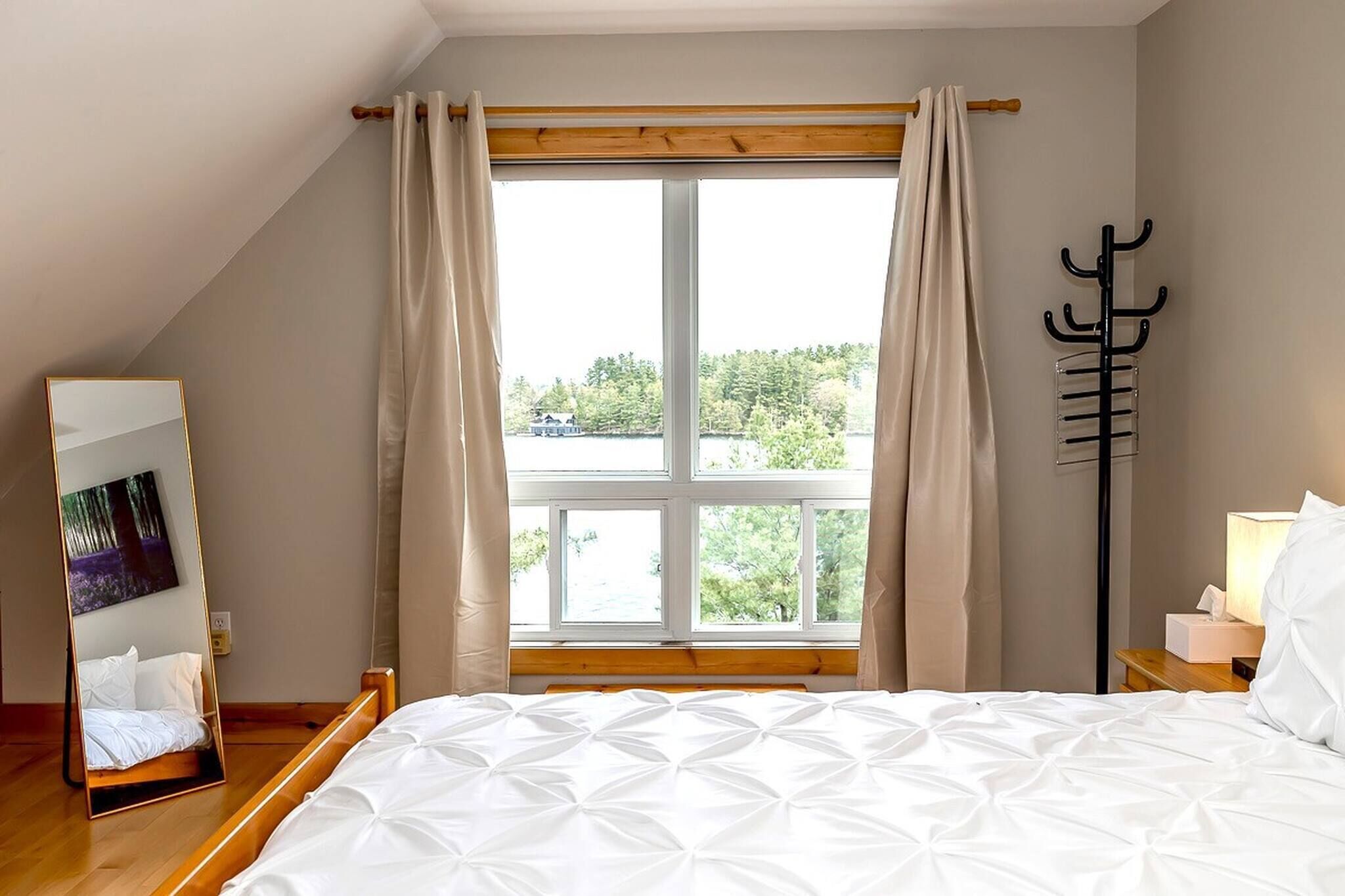 A bedroom with a large window, a bed, a bedside table with a lamp, and a coat rack.