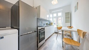 A modern kitchen with stainless steel appliances, a white dining area with a wooden chair, and a window providing natural light. - Apartament Przytulny na Starym Mieście 2 sypialnie (Gdansk)