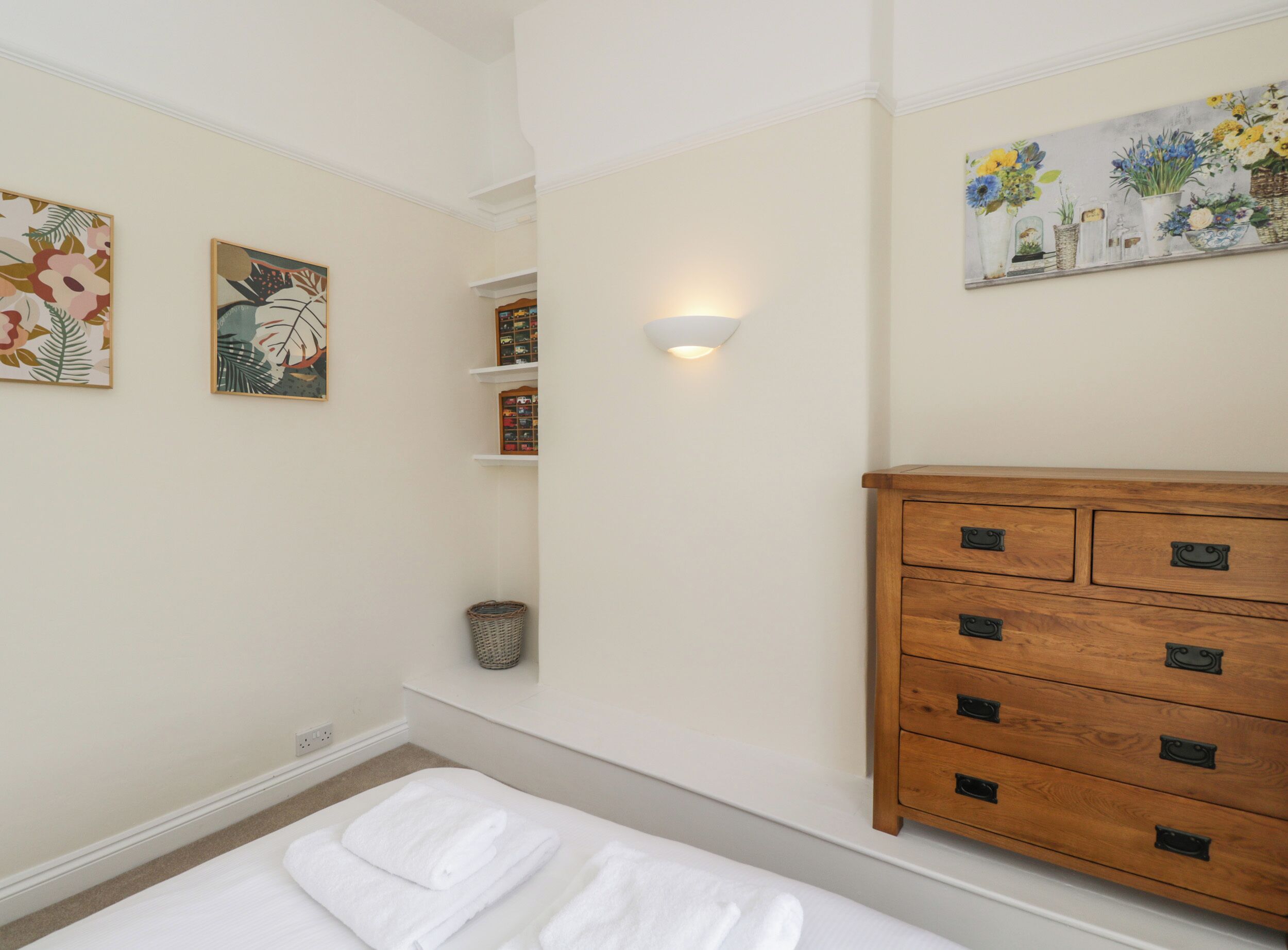 Ein Schlafzimmer mit einem Bett, einer hölzernen Kommode, Wandregalen und zwei gerahmten Kunstwerken.