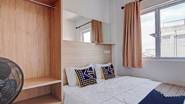 Une chambre à coucher avec un lit, un ventilateur, une fenêtre avec des rideaux et une vue sur un bâtiment à l’extérieur.