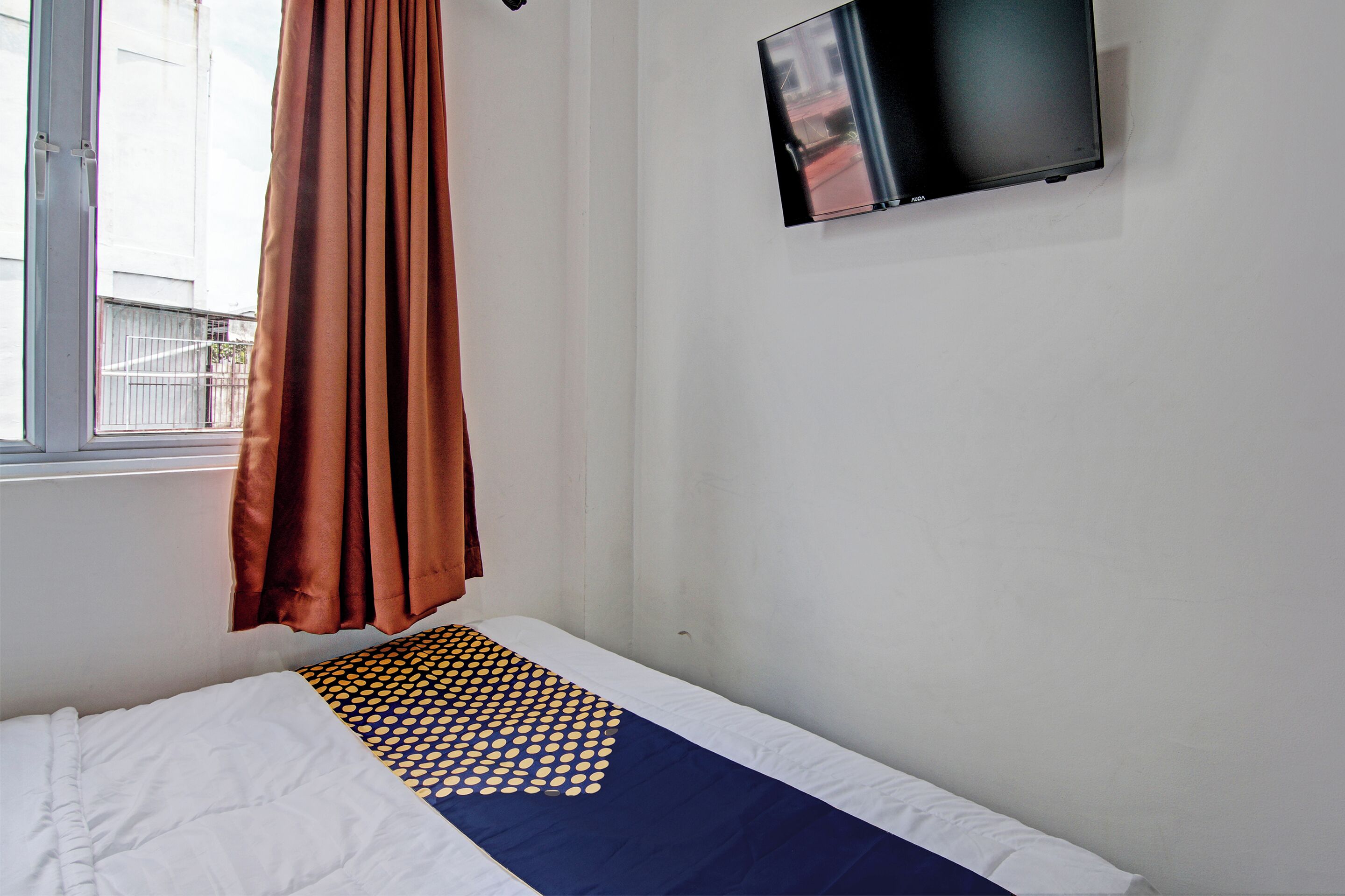 A single bed with a patterned bedspread, a window with curtains, and a flat-screen TV mounted on the wall.
