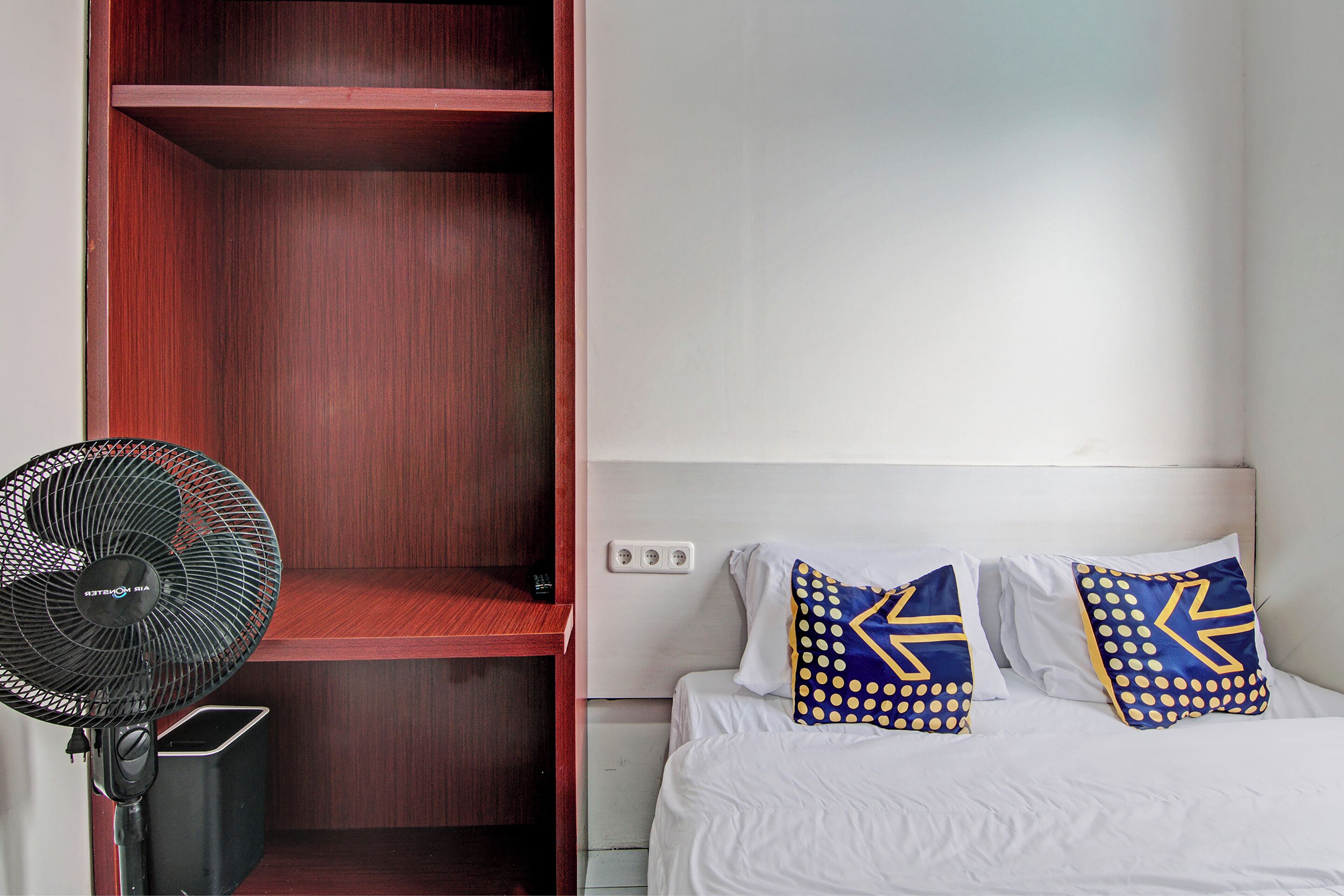 Quarto de hotel com cama, duas almofadas com padrão geométrico, ventilador e guarda-roupa de madeira.