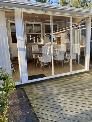 A covered patio with a table and chairs, a wooden deck, and a glass door leading into a room with a bed and a window.