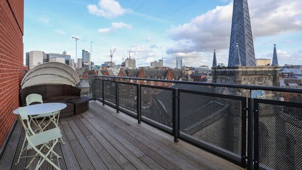 A rooftop terrace with a wooden deck, a white table and chairs, and a view of the city skyline including a tall building. - 2 Bed Duplex close to Millennium Sq (Leeds)