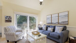 Ein Wohnzimmer mit einem gestreiften Sessel, einem blauen Sofa, einem Glastisch und Blick ins Freie durch eine Glasschiebetür.