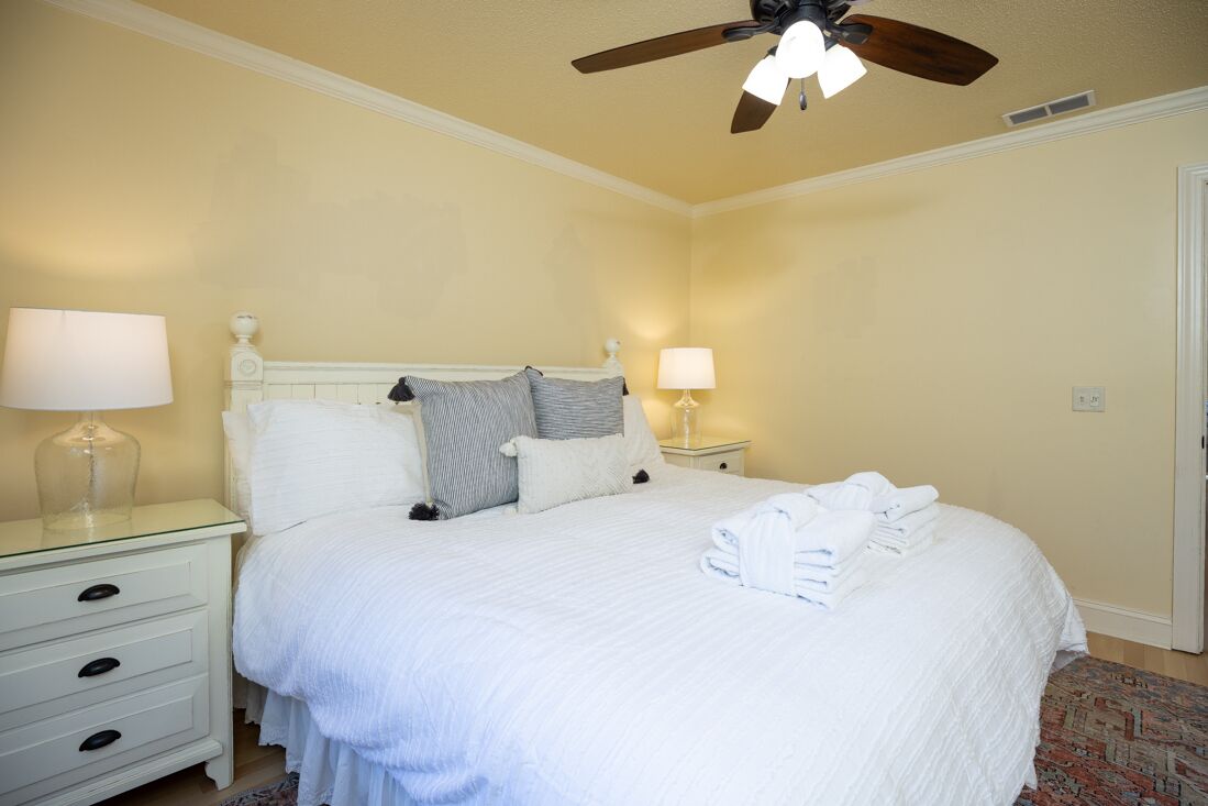 Un lit bien fait, avec du linge de lit blanc et une serviette pliée posée sur le côté. On peut voir deux tables de chevet avec des lampes, un ventilateur de plafond et une porte au fond.