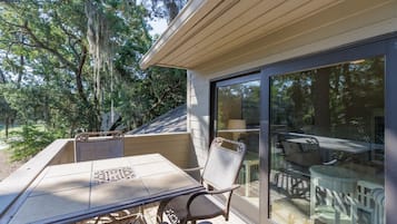 Un patio exterior con una mesa y sillas, un ventanal de vidrio que da a una terraza, y árboles circundantes.