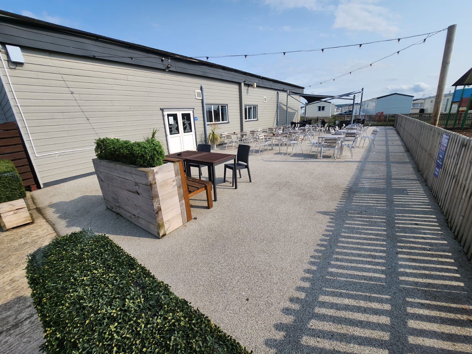 Een terras met buitenmeubels, een houten tafel en stoelen, en een gebouw op de achtergrond.