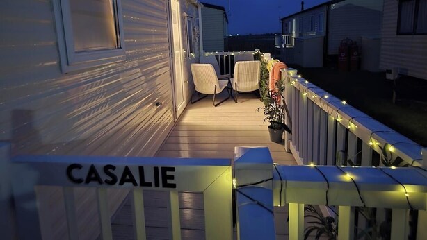 Una caravana con terraza, un letrero 'CASALIE', mobiliario exterior y barandilla con luces.