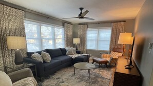 A living room with a sectional sofa, a round coffee table, a side table, a lamp, a ceiling fan, and a view through large windows. - The Warm & Inviting Place (Sioux Falls)