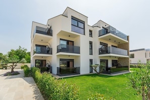 A modern multi-story residential building with balconies, a well-maintained lawn, and a clear sky.