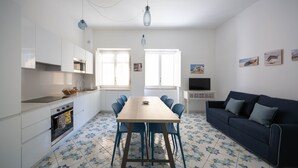 Una cocina moderna con armarios blancos, una mesa de comedor de madera y sillas azules. Hay un sofá azul oscuro y un televisor sobre un mueble.