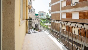 Un balcón estrecho con barandilla de metal, suelo de baldosas y vistas a edificios cercanos.