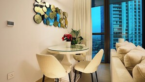 Una sala de estar moderna con una mesa redonda de cristal, dos sillas, un sofá, una pared decorativa con espejos y vistas a un paisaje urbano a través de amplios ventanales.