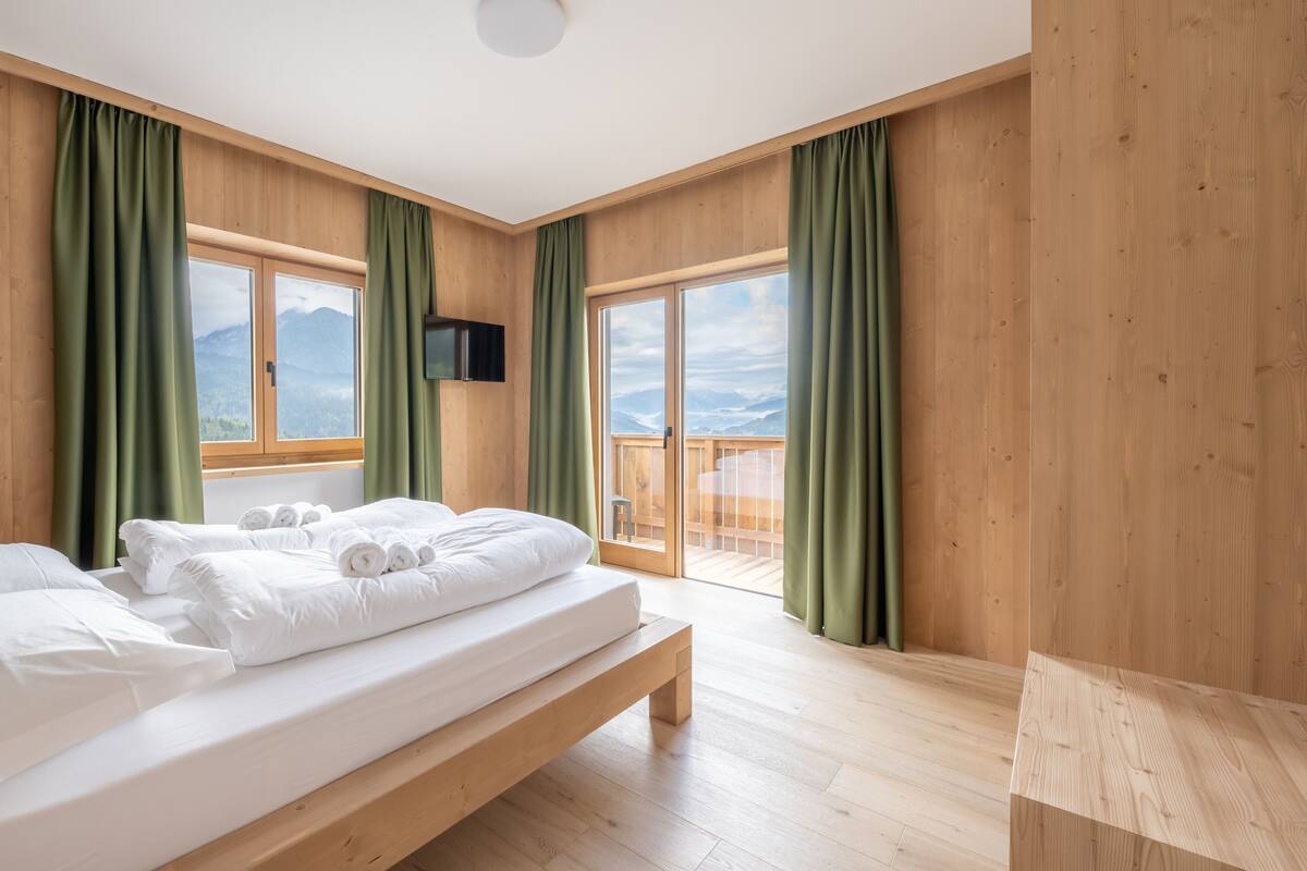Une chambre moderne avec un plancher en bois, un grand lit recouvert de draps blancs, des rideaux verts et un téléviseur fixé au mur.