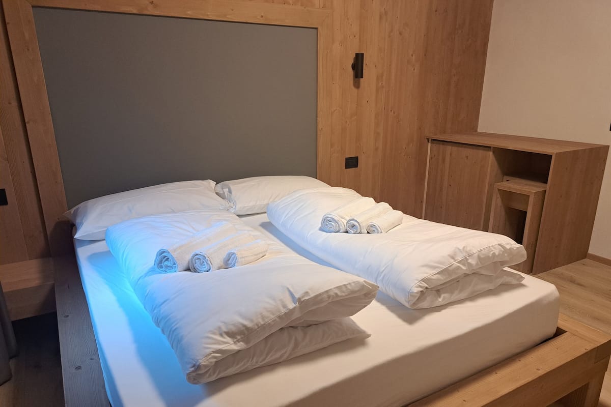 Un lit bien fait, avec du linge de lit blanc et des serviettes pliées, une tête de lit en bois et une petite table de chevet en bois.