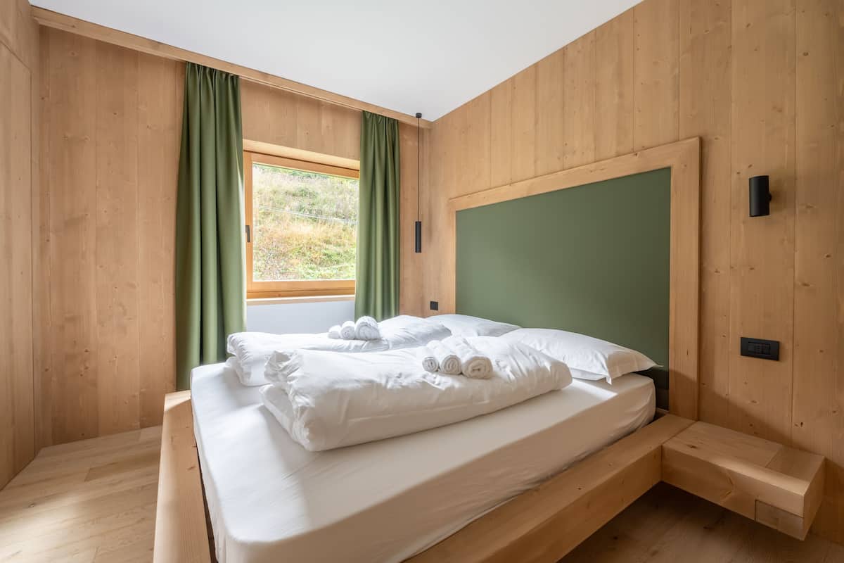 Une pièce aux murs lambrissés, avec un lit, des rideaux verts et une vue sur la verdure à l’extérieur.