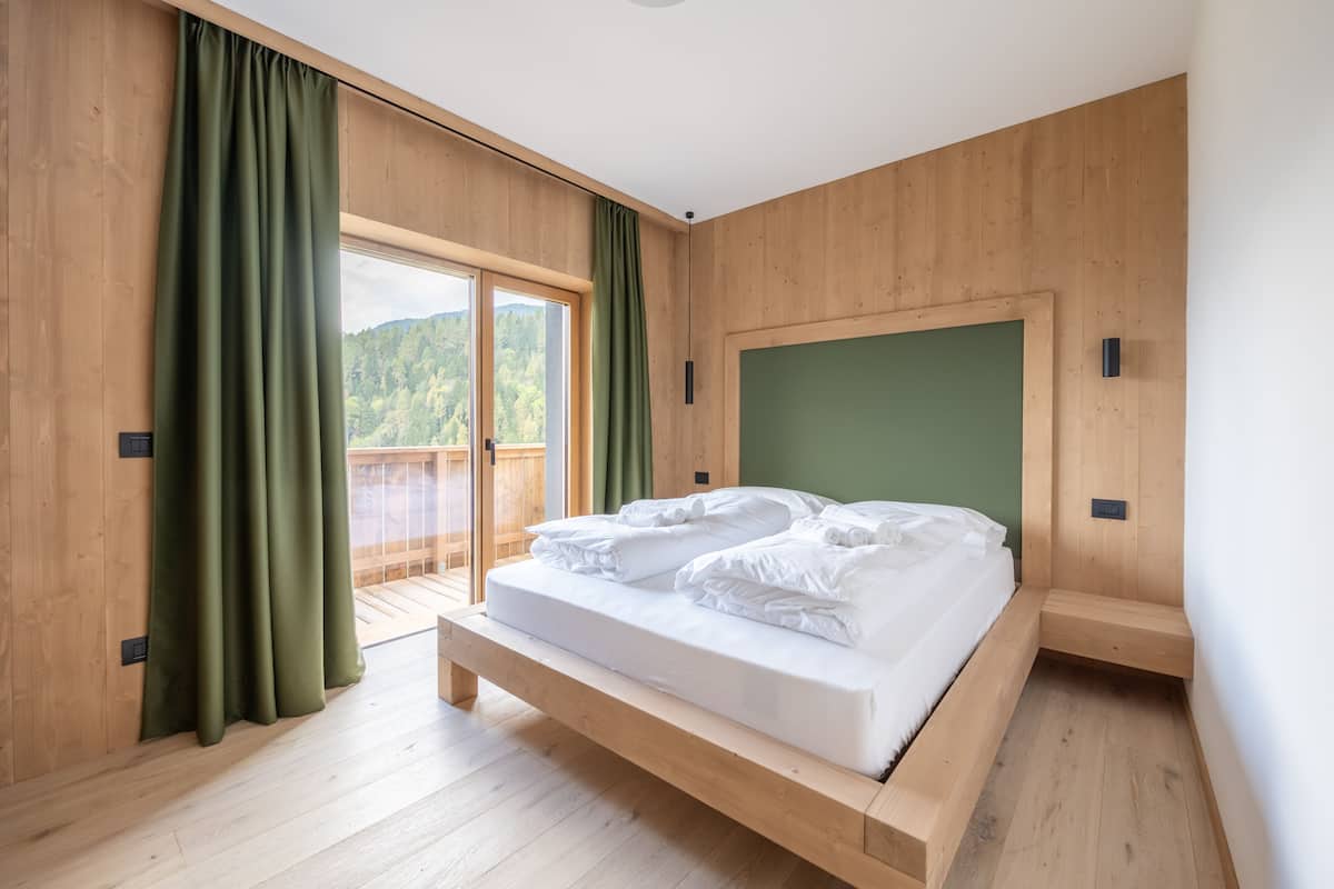 Une chambre aux murs lambrissés, avec un lit, des rideaux verts et une vue sur une zone boisée.