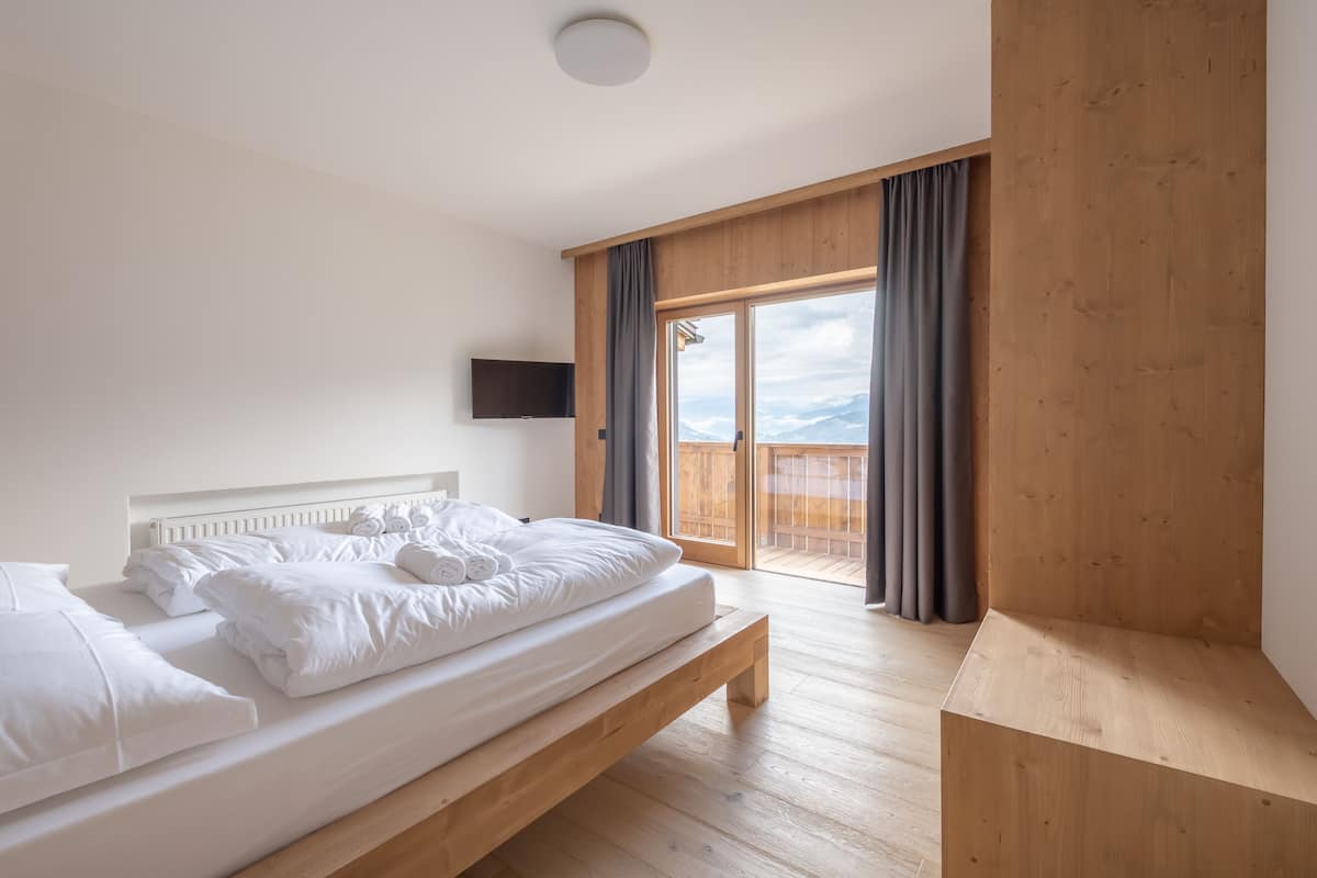 Une chambre moderne avec un lit en bois, un téléviseur fixé au mur et un balcon offrant une vue sur les montagnes.