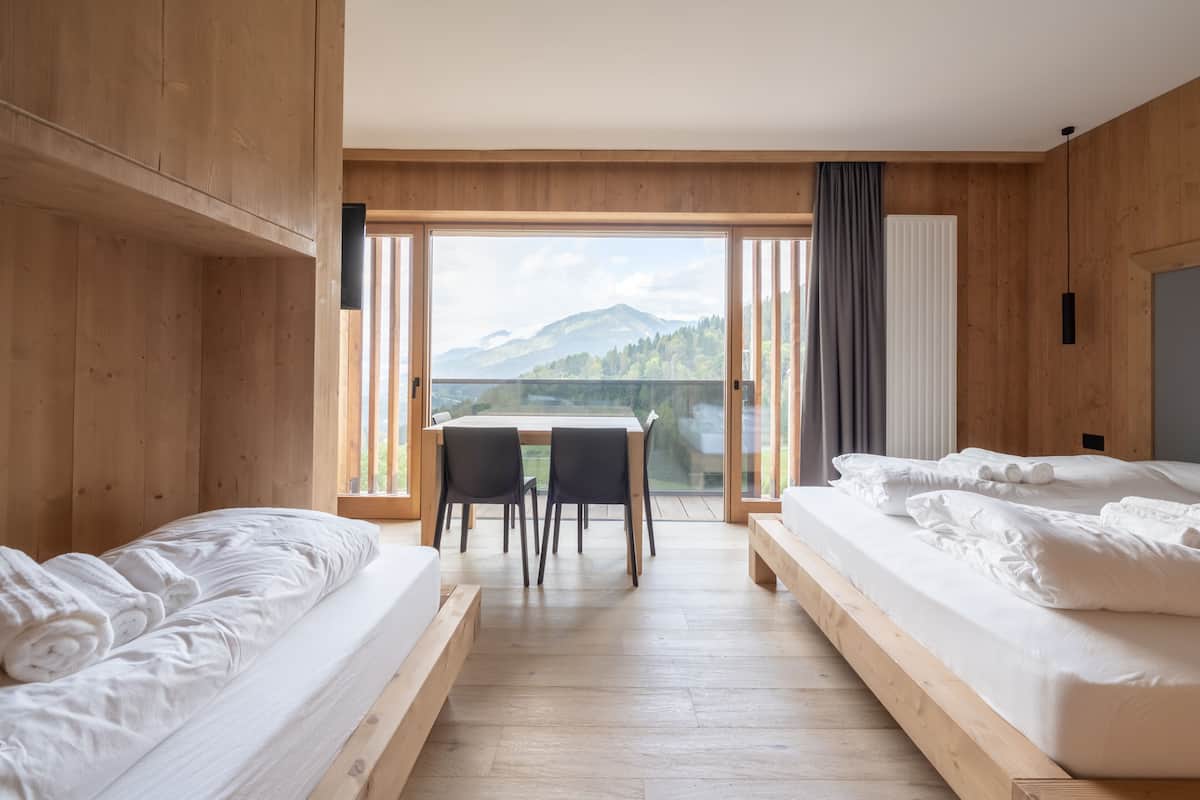 Une chambre avec deux lits, un plancher en bois, une table avec des chaises et une vue sur les montagnes, visible à travers de grandes fenêtres.