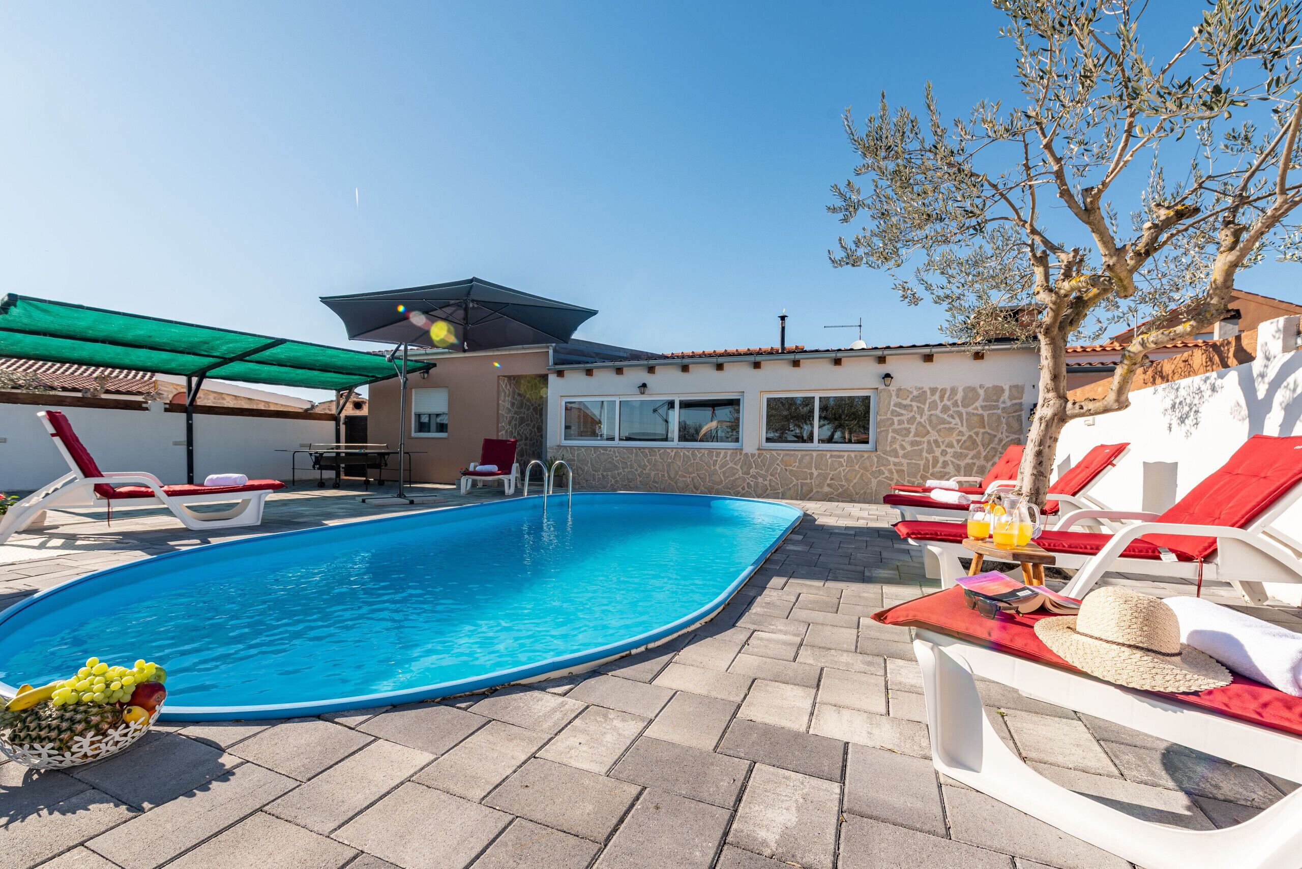 Una piscina con sillones de descanso y una mesa con bebidas, rodeada por un muro de piedra y un edificio con ventanas grandes.