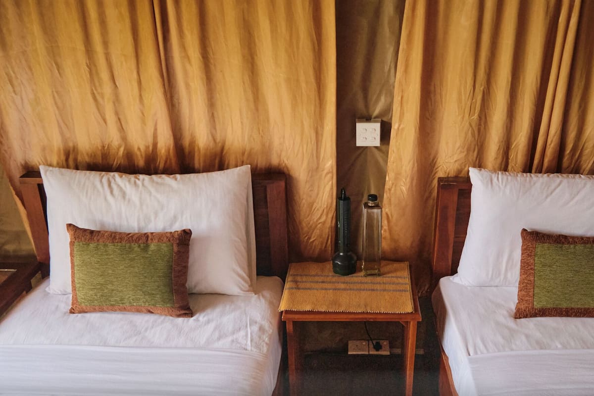 A room with two single beds, each with a white pillow and a green accent pillow. There is a small wooden table between the beds with a black bottle and a clear bottle on it.