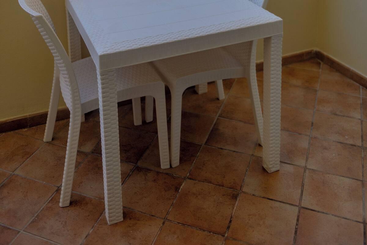 Une table et des chaises en osier blanc sont disposées sur un balcon dont le sol est carrelé, offrant une vue sur un immeuble et des arbres.