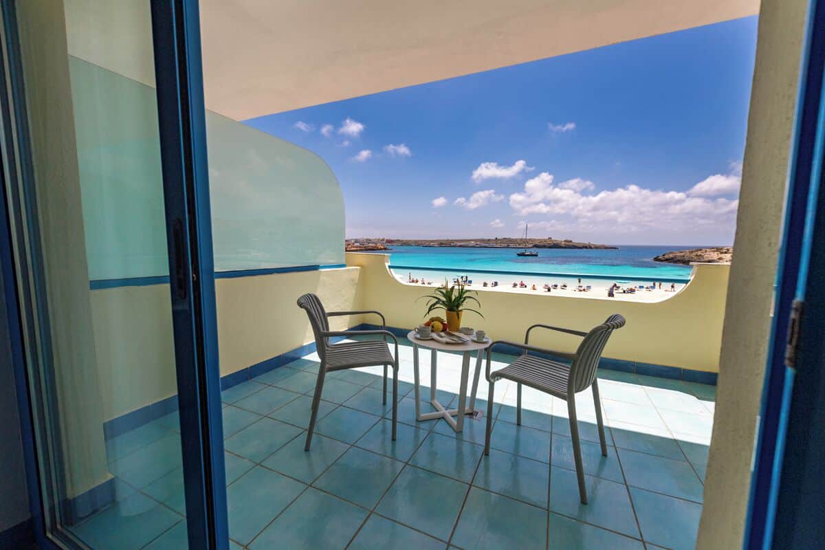Un balcon avec deux chaises et une table, donnant sur une plage aux eaux bleu clair et sur laquelle se trouve un bateau.