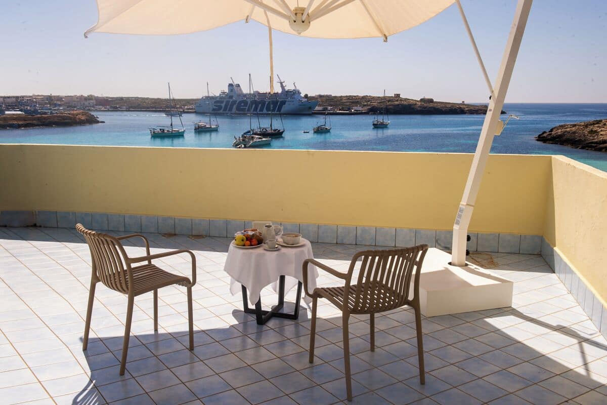 Un balcon avec deux fauteuils en osier et une petite table recouverte d’une nappe blanche, un navire nommé « siremur » en arrière-plan, et une vue dégagée sur la mer et le littoral lointain.