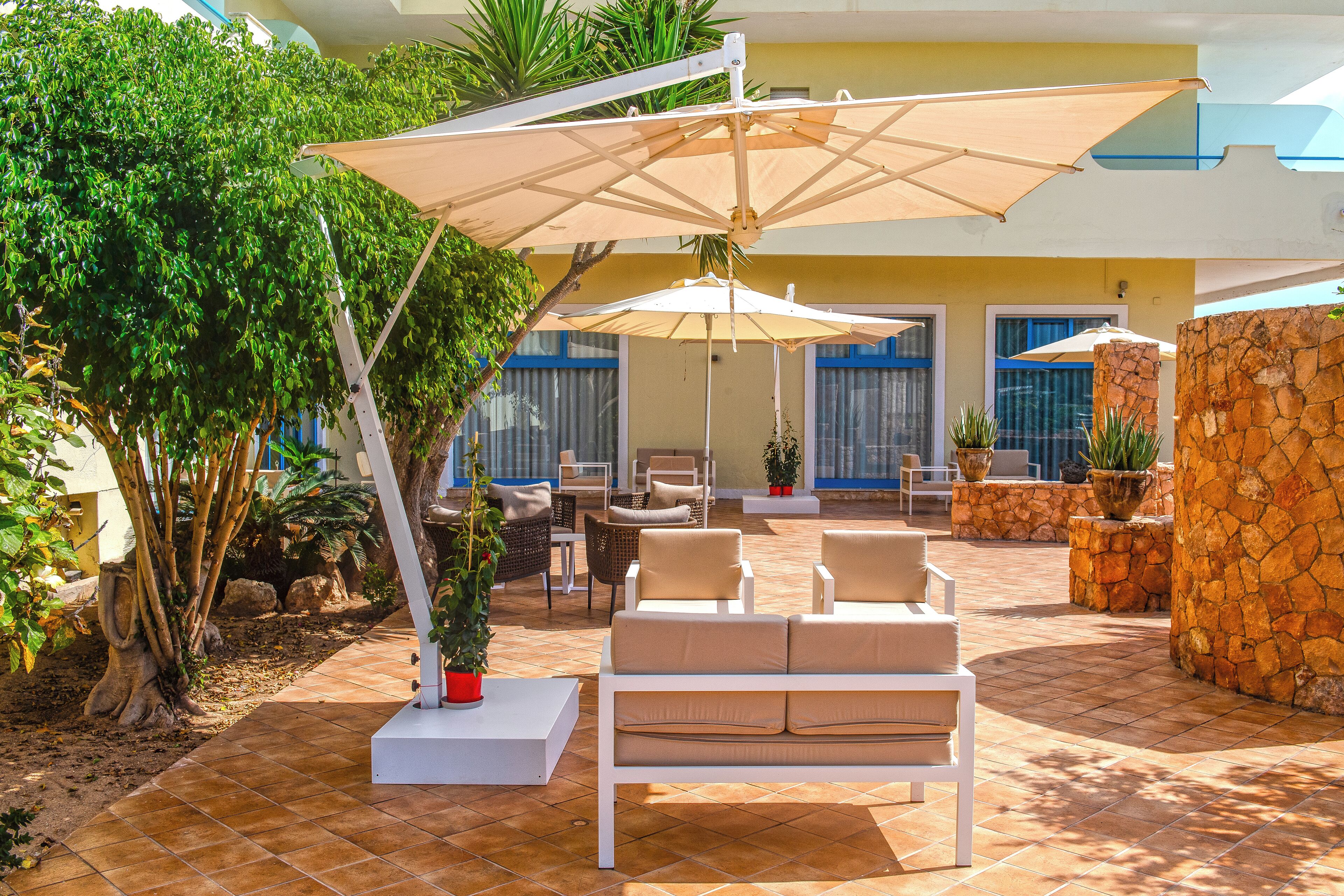 An outdoor patio area with a white sofa, beige cushions, and a large umbrella providing shade. There are potted plants and a stone wall in the background.