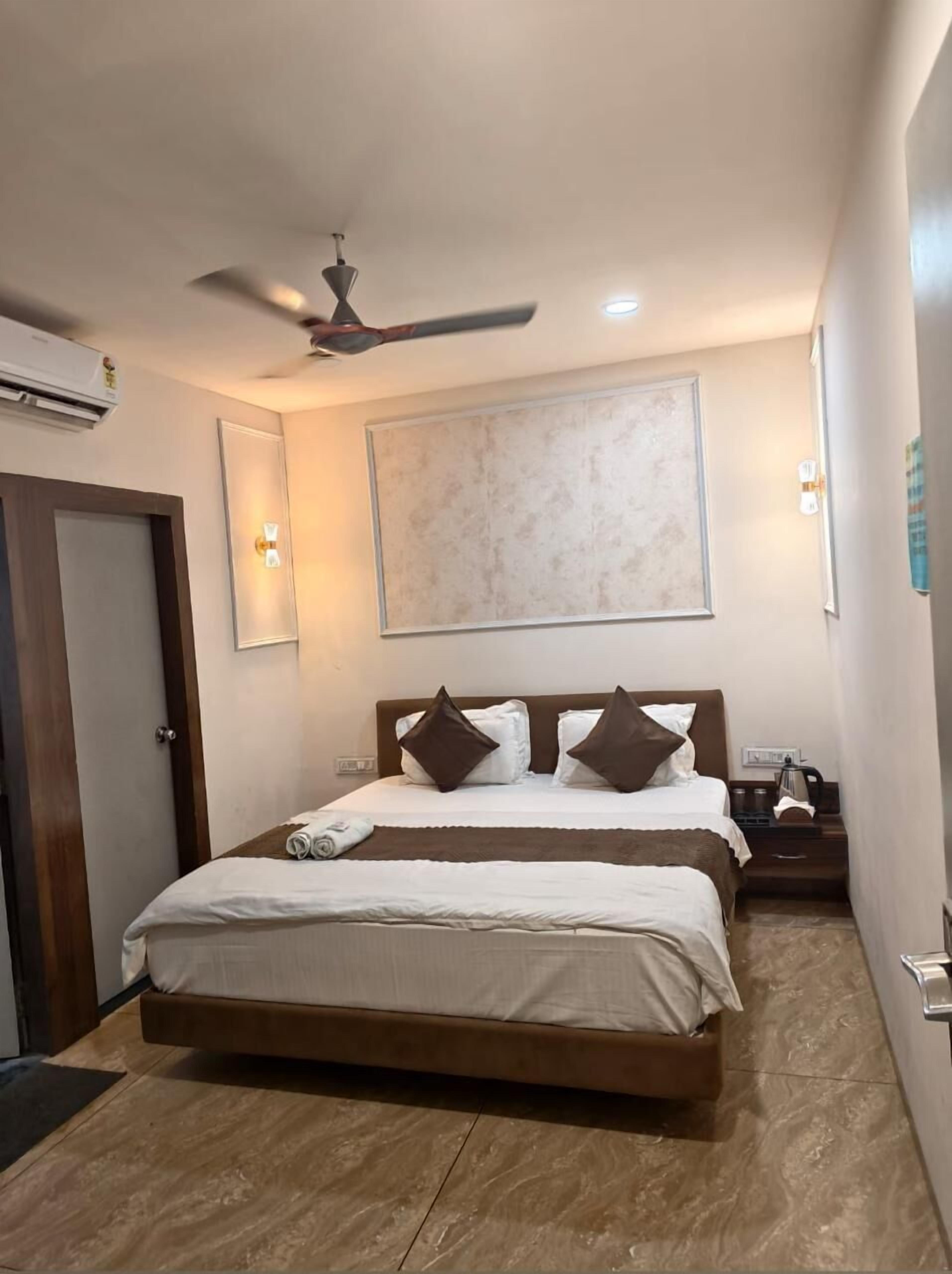 A hotel room with a bed, a ceiling fan, a wall-mounted air conditioner, and a nightstand with a lamp.