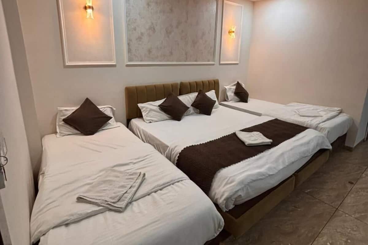 Quarto de hotel com três camas, cada uma com lençóis brancos e almofadas castanhas. Aparelhos de luz fixados na parede e um ventilador de teto.