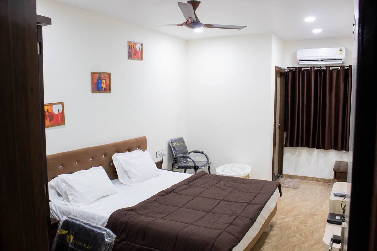 Quarto com cama, uma cadeira, mesa pequena, ar-condicionado e ventilador de teto.