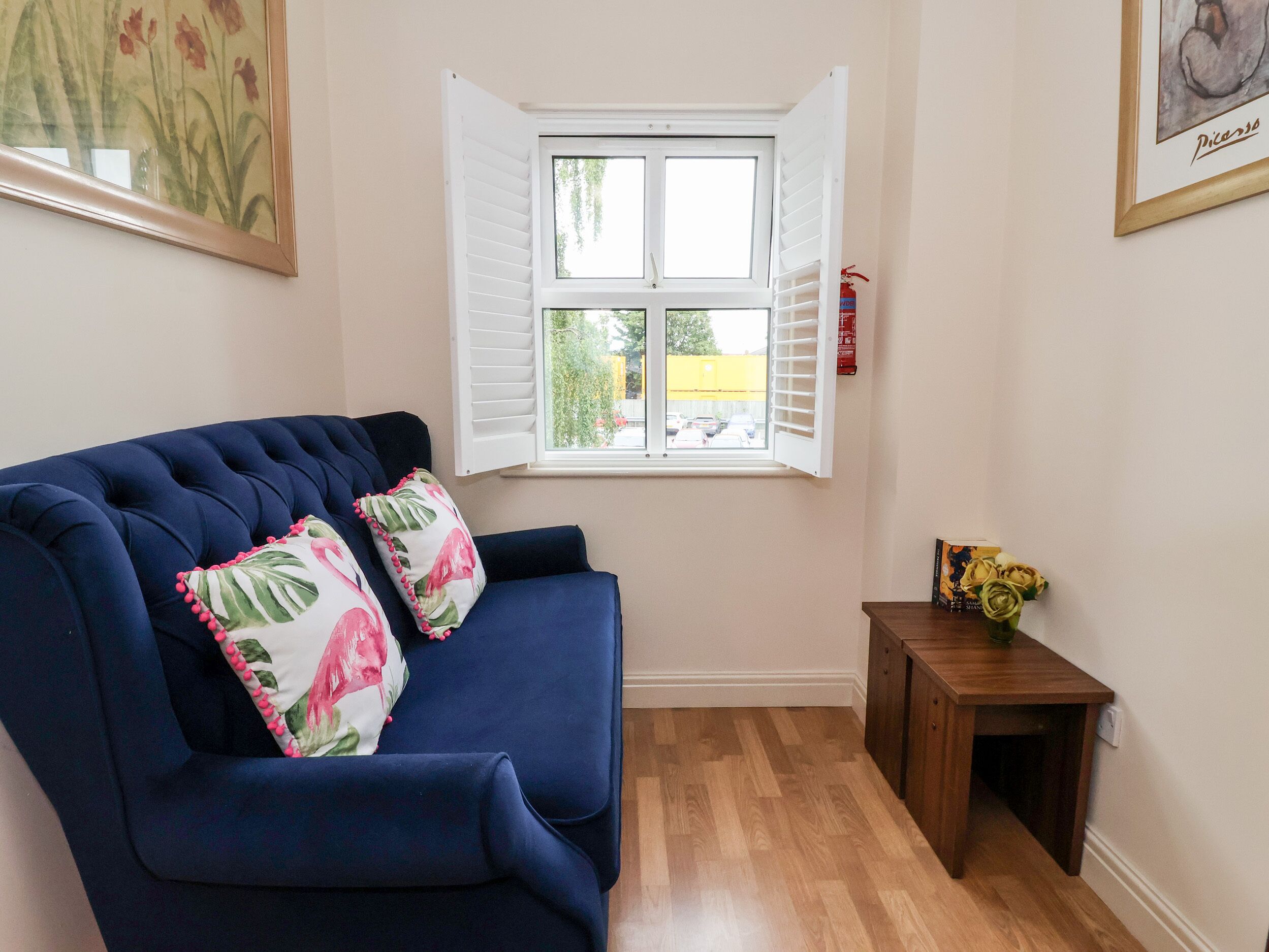 Ein Wohnzimmer mit einem dunkelblauen Sofa, zwei floralen Kissen, einem kleinen Holznachttisch mit einer Blumenvase und einem Fenster mit weißen Fensterläden.