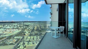 Un balcon doté d’une balustrade en verre, d’une table et de deux chaises, offrant une vue sur un paysage urbain avec ses bâtiments et ses rues.
