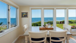 A dining area with a table and chairs, large windows with a view of the sea, and a framed picture on the wall. - Sea Spray - Beautiful 2BR, 1BA cottage with expansive views of Casco Bay (Bailey Island)