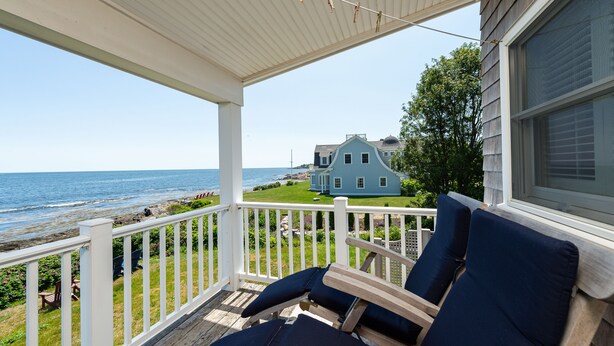 A covered porch with two blue cushioned chairs, a white railing, and a view of the ocean and a house in the background. - Sea Spray - Beautiful 2BR, 1BA cottage with expansive views of Casco Bay (Bailey Island)