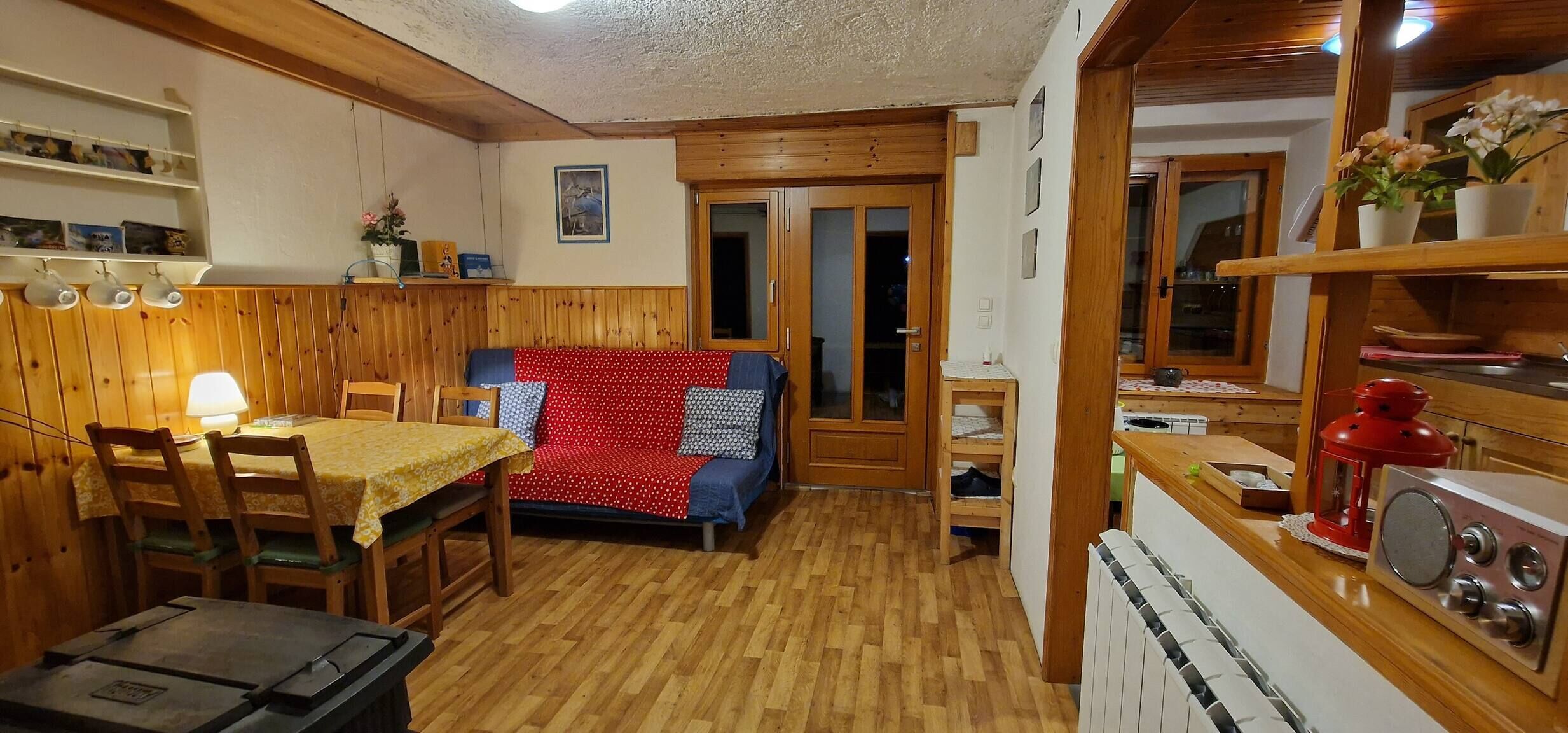 Ein gemütliches Zimmer mit Holzboden, einer Couch mit roten und blauen Punkten, einem Esstisch mit Stühlen und einer Küchenzeile mit Holzschränken und einem roten Gerät.