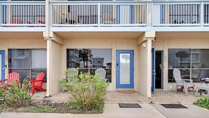 A two-story building with a beige facade, blue-framed windows, and a front entrance with a blue door. There are red and white Adirondack chairs outside, and a small garden area with plants. - Relax Inn - Seamist 156 (Port Aransas)