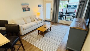 A living room with a sofa, coffee table, and a dining area with chairs and a table. There is a ceiling fan, a window with curtains, and a view of the outside. - Relax Inn - Seamist 156 (Port Aransas)